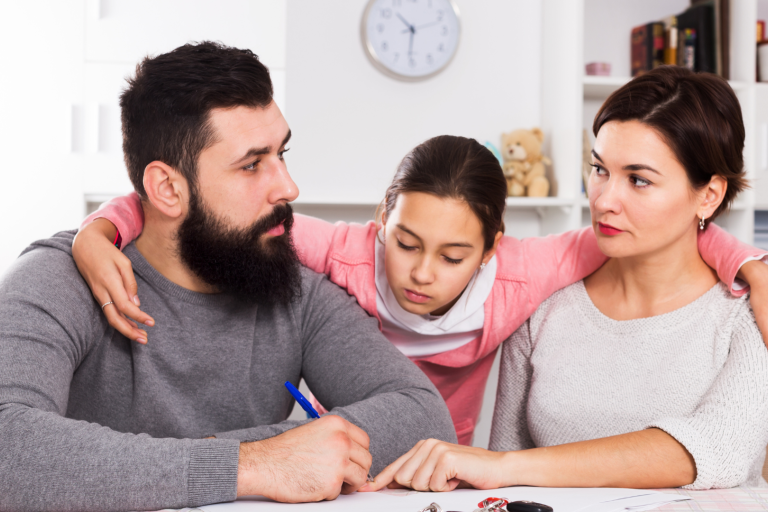 Parents sitting at table looking at paperwork with teenage girl with her arms wrapped around both parents