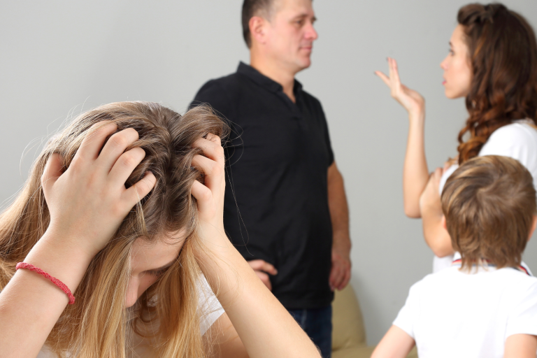 Teenager with head in hands and parents arguing in background with infant looking on